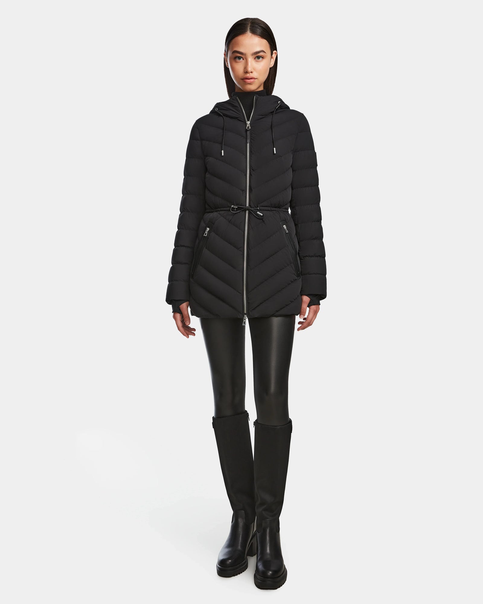 A woman stands facing forward against a plain white background, wearing the Rudsak FRANCINE - 8124722 Black quilted puffer jacket with a belt, paired with black leather-look pants and knee-high black boots.