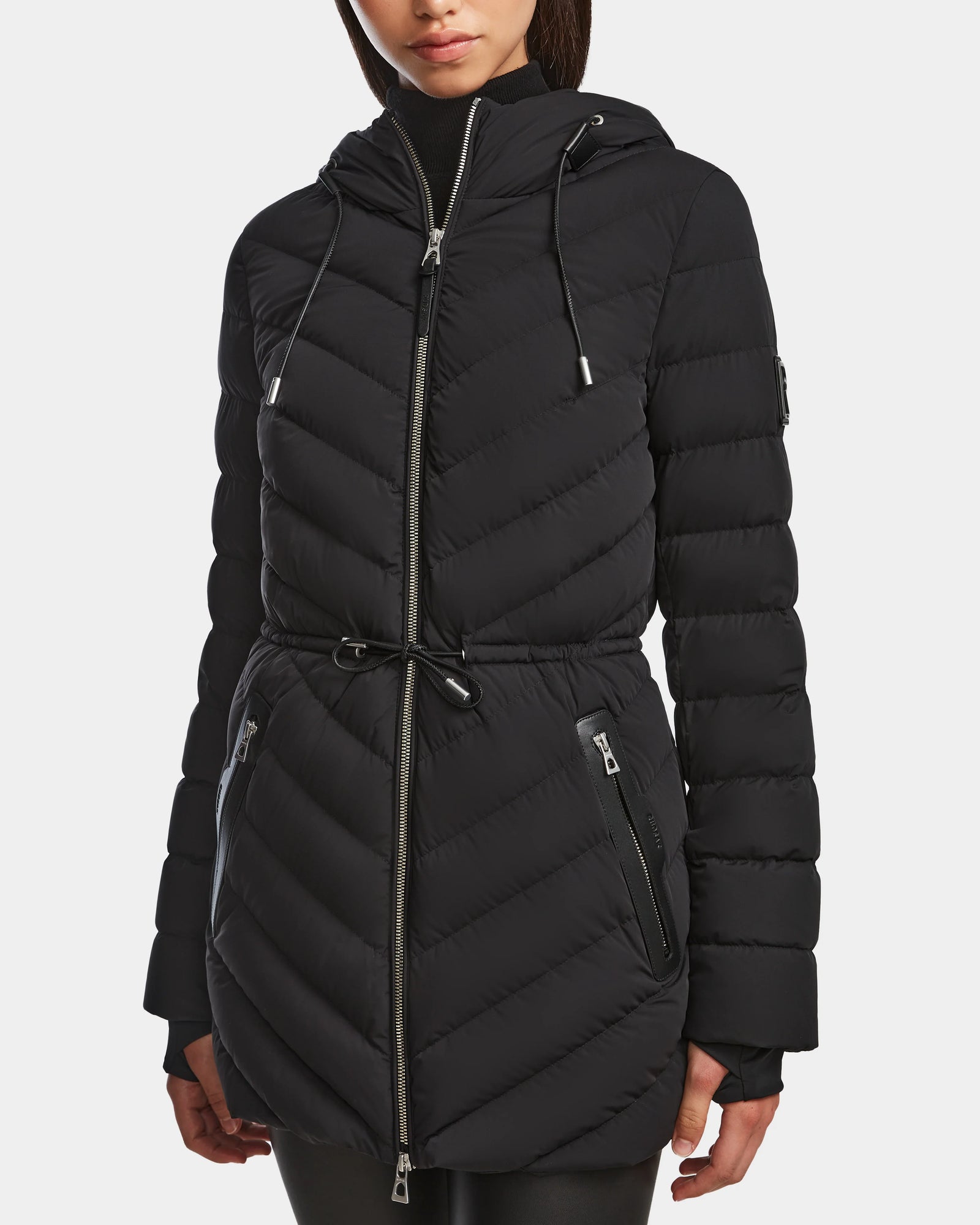 A person wears the Rudsak FRANCINE - 8124722 Black quilted puffer jacket with a drawstring waist, zippered front, and pockets against a plain white background—this stylish women’s light down puffer is ideal for chilly days.