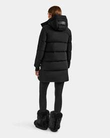 A person wearing the Rudsak ALESSANDRA M - L125552 Black women's down puffer, black leggings, and black furry boots stands with their back to the camera against a plain light background.