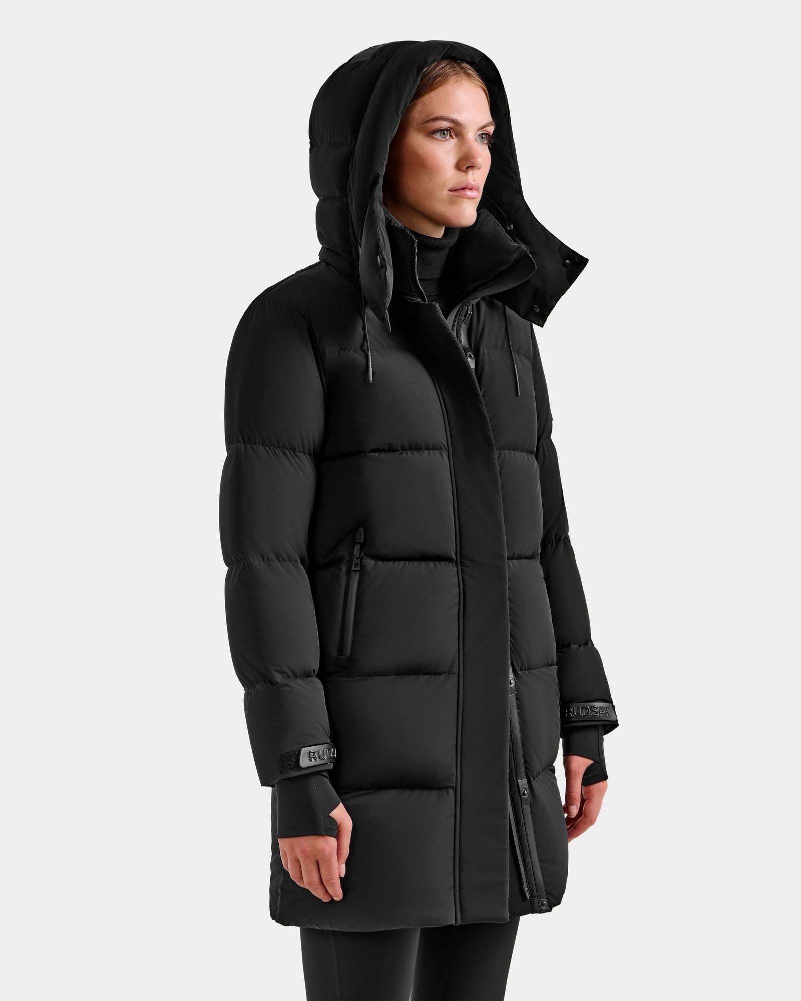 A woman wears the Rudsak ALESSANDRA M - L125552 Black down puffer coat with a hood and high collar, featuring zippered pockets and visible stitching, standing against a plain light gray background.