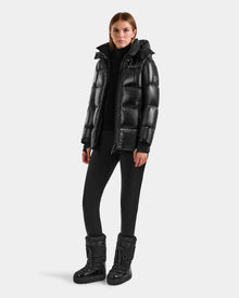 A woman wearing the Rudsak ALESSANDRA S-S - L125560 Black down puffer with a black turtleneck, leggings, and winter boots stands against a plain light gray background.