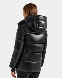 A person with straight, light brown hair wears the Rudsak ALESSANDRA S-S - L125560 Black glossy puffer jacket with hood. The Rudsak logo is visible on the back and left sleeve. The background is plain and light-colored.