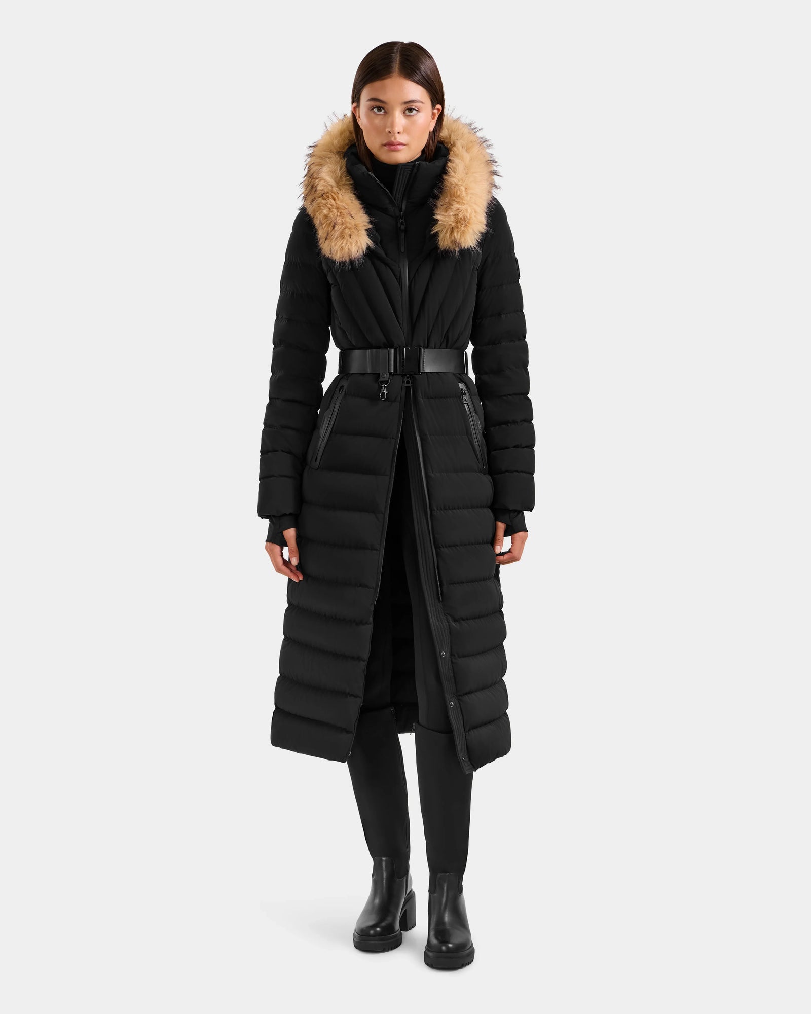 A woman with straight hair wears the Rudsak ALISA L - L125574 Black belted, quilted puffer coat with RDS-certified down and a tan faux-fur trim hood, paired with black pants and ankle boots against a plain white background.