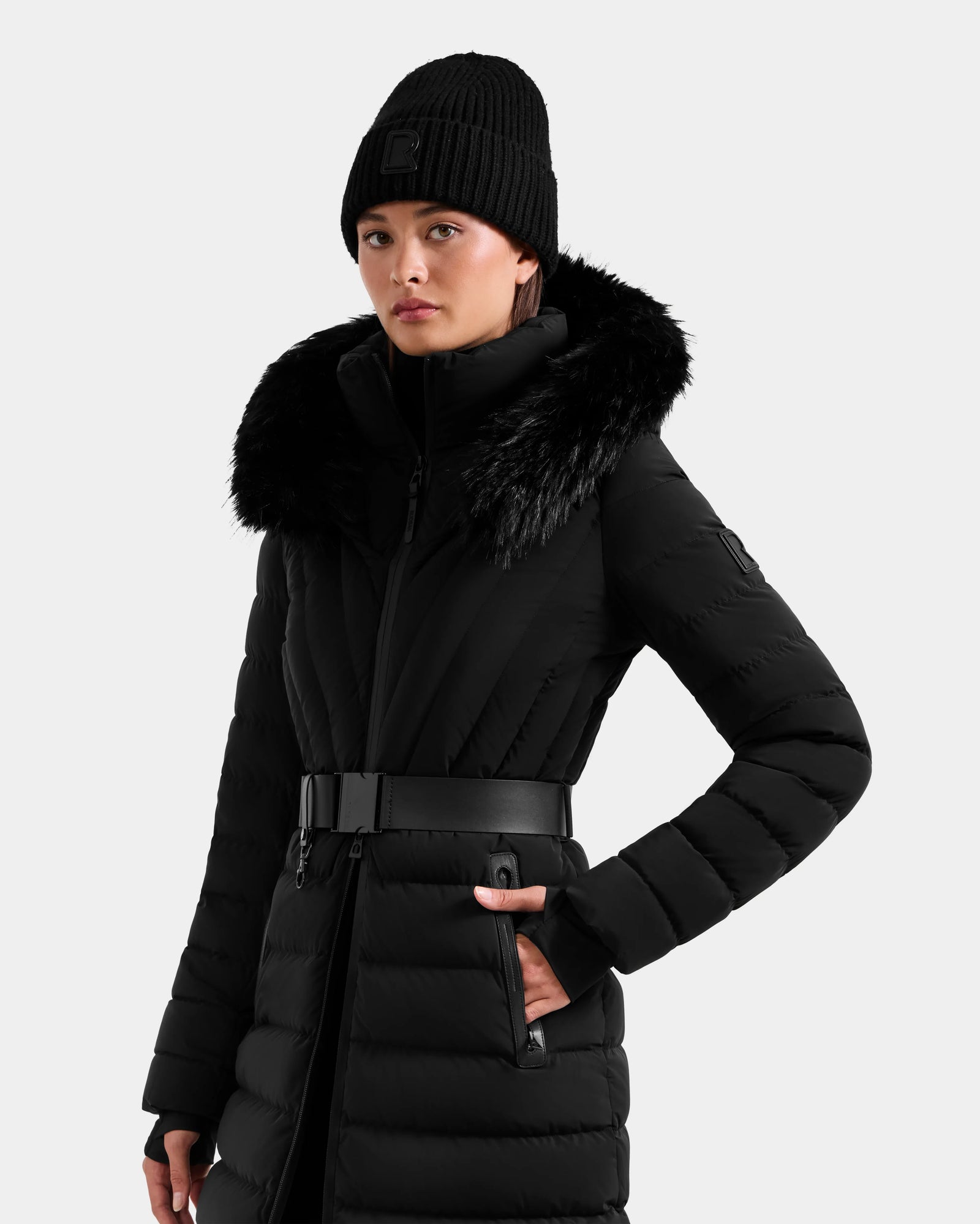 A person wears the Rudsak ALISA M - L125586 Black quilted down parka with faux fur-trimmed hood, RDS-certified fill, and vegan leather belt, paired with a black knit beanie, against a plain light background.