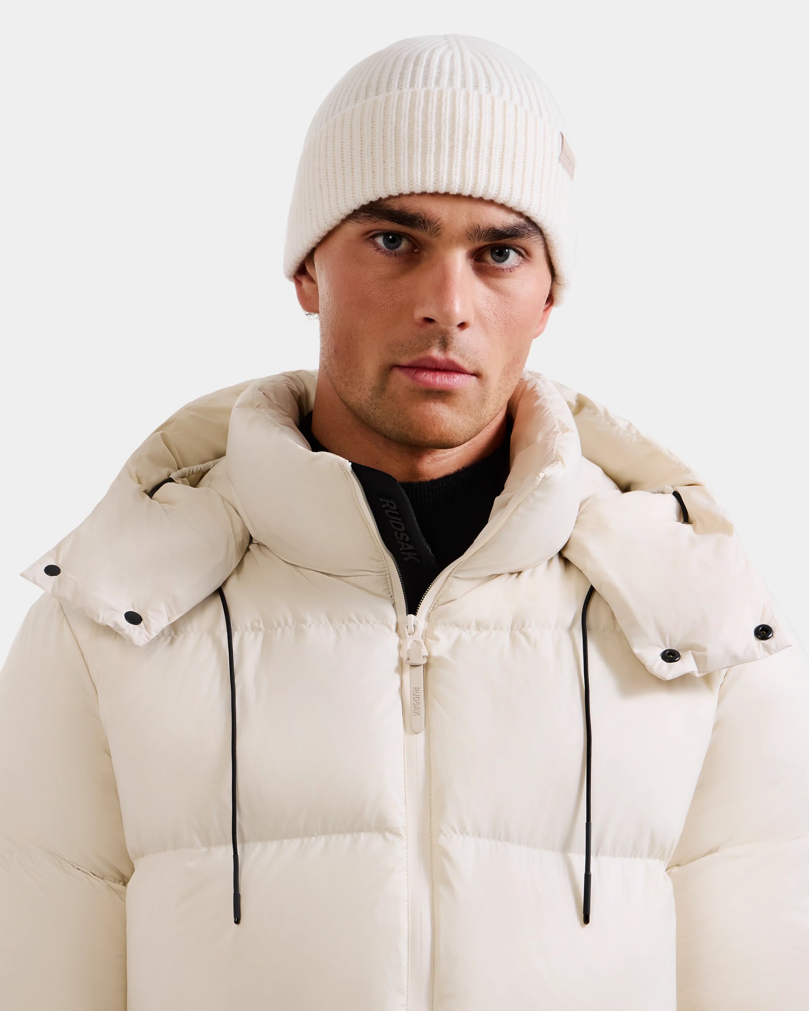 A man wears Rudsak's ANDREY - U425507 Vanilla white puffer jacket with a hood and a wool-cashmere beanie, looking directly at the camera against a plain light background.