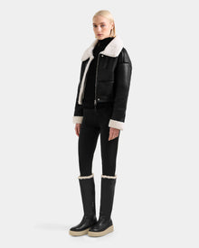 A woman wears the Rudsak APRIL - L125517 Black shearling aviator jacket with a white fur collar and cuffs, paired with a black turtleneck, leggings, and black knee-high boots featuring white trim and beige soles against a plain background.