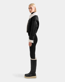 A woman with blonde hair stands in profile against a light gray background, wearing the Rudsak APRIL - L125517 Black shearling jacket, paired with black leggings and tall black platform shearling boots.