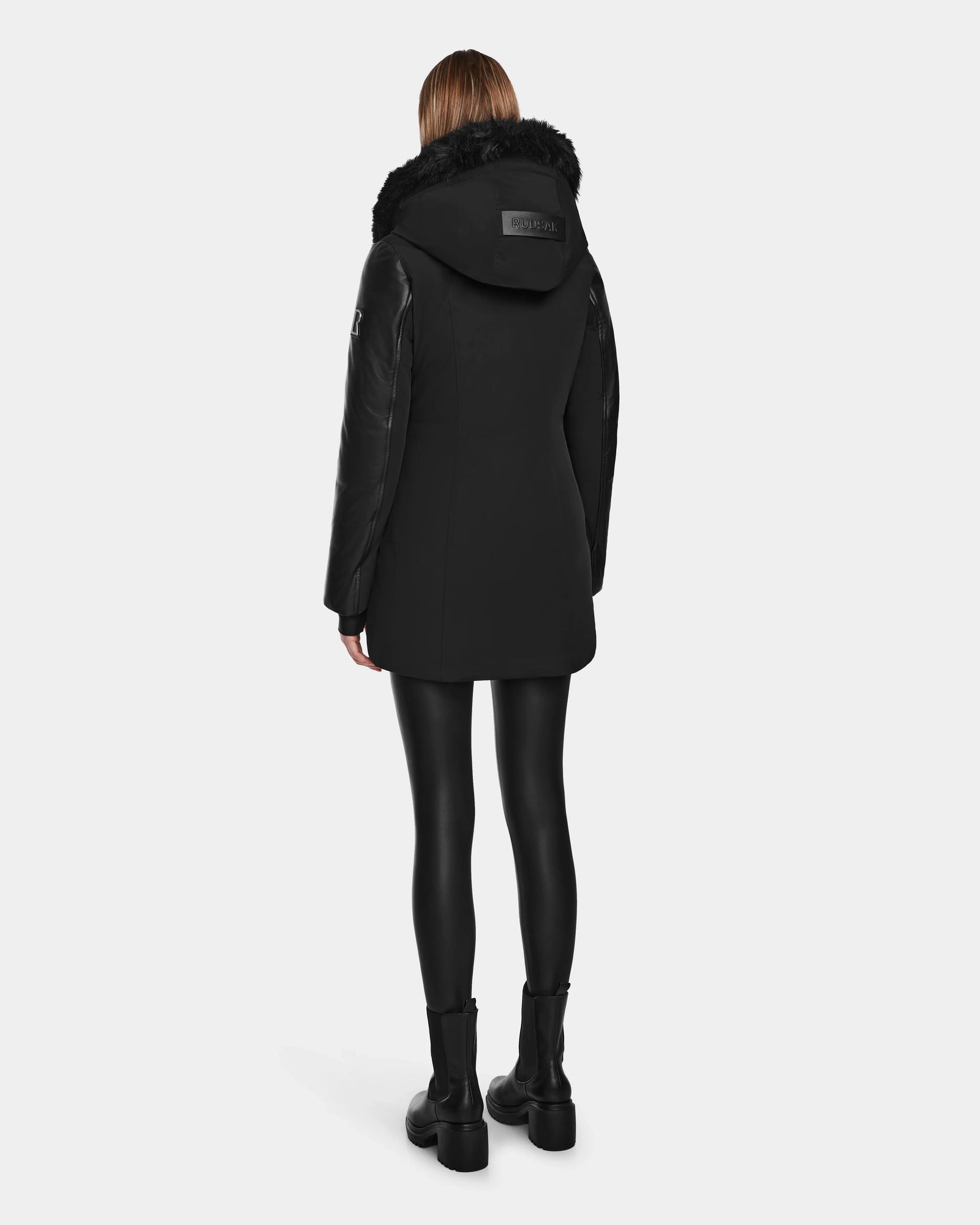 A person faces away, wearing the Rudsak AURELLE - L124538 Black women’s down parka with vegan leather sleeves and fur-trimmed hood, paired with black leggings and boots—offering stylish winter protection against a light background.