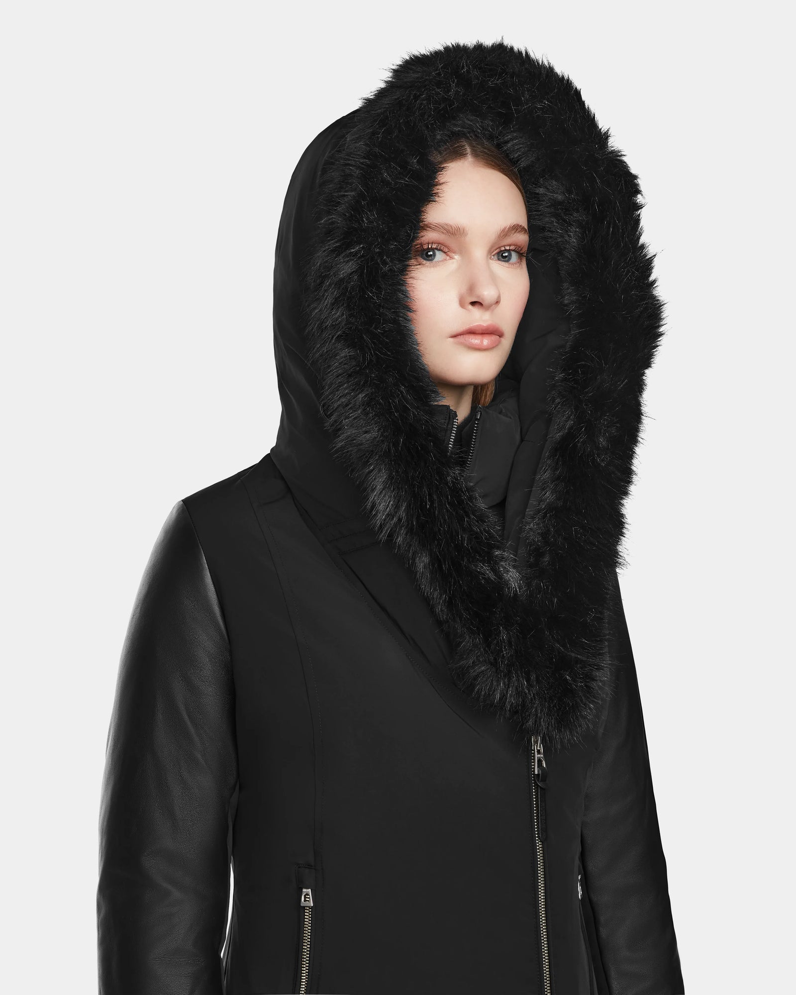 A woman models the Rudsak AURELLE - L124538 Black down parka, featuring a large faux fur hood and vegan leather sleeves, as she looks at the camera against a plain light background.