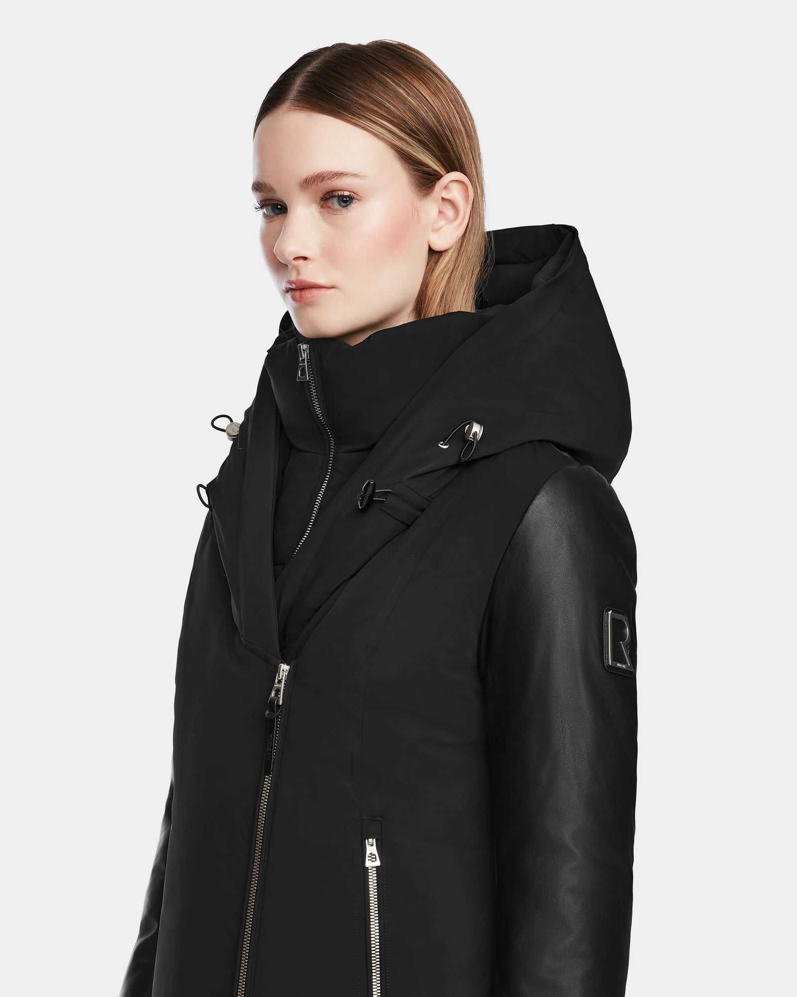 A woman with straight, light brown hair models the Rudsak AURELLE - L124538 Black coat, featuring vegan leather sleeves and silver zippers, against a plain light background.