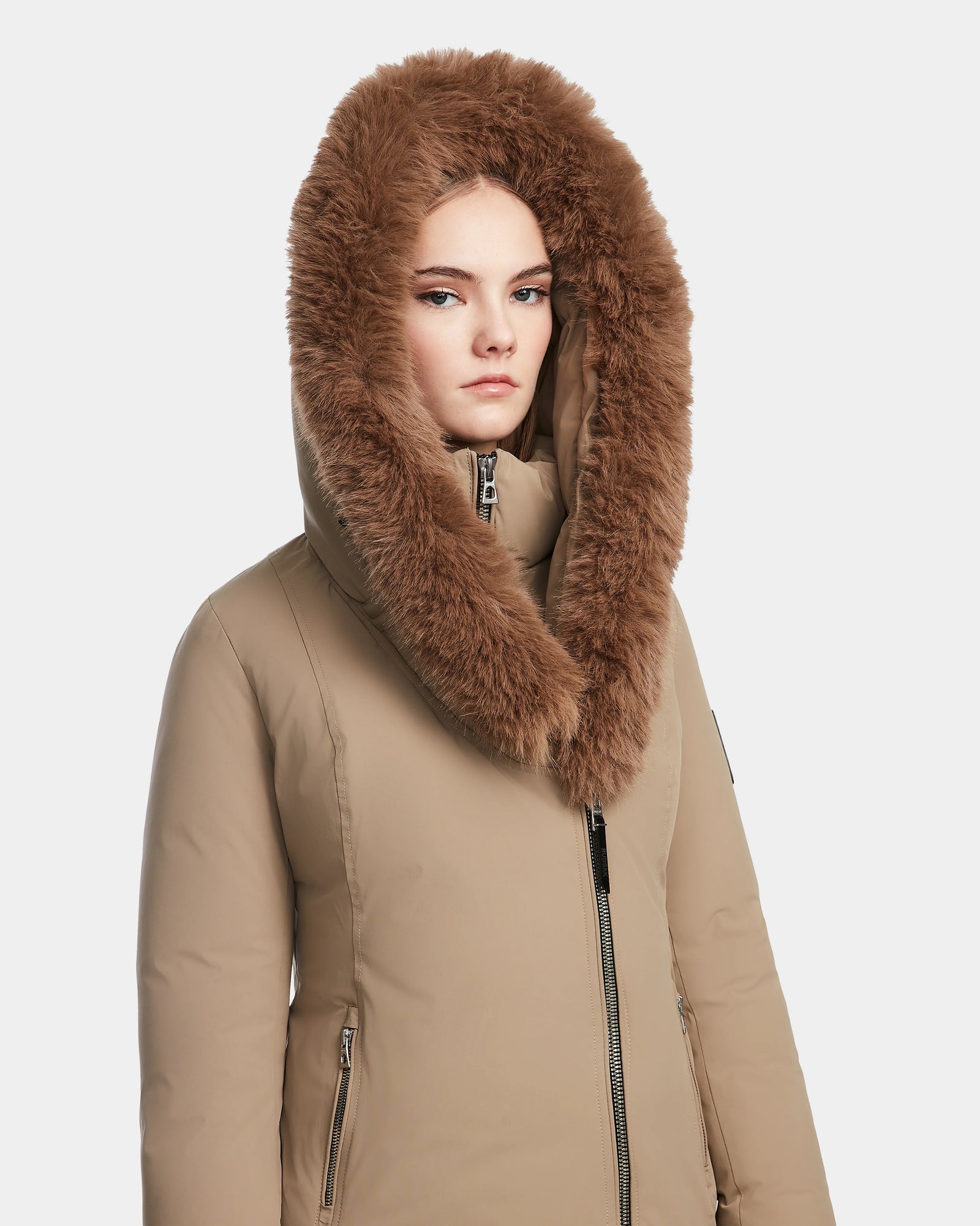 A woman models the Rudsak AURORE - L123561 Sand fitted winter coat with a large, fluffy brown fur-lined hood, looking directly at the camera against a plain white background.