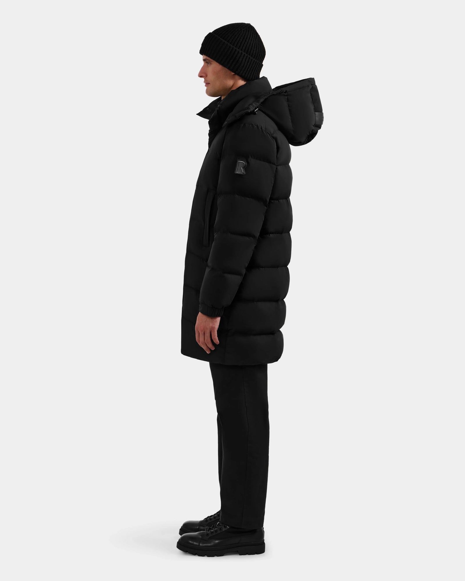 A man stands in profile against a white background wearing the Rudsak BRAYDEN M-M - M125535 Black weather-resistant men’s down puffer with hood, paired with a black beanie, pants, and boots.