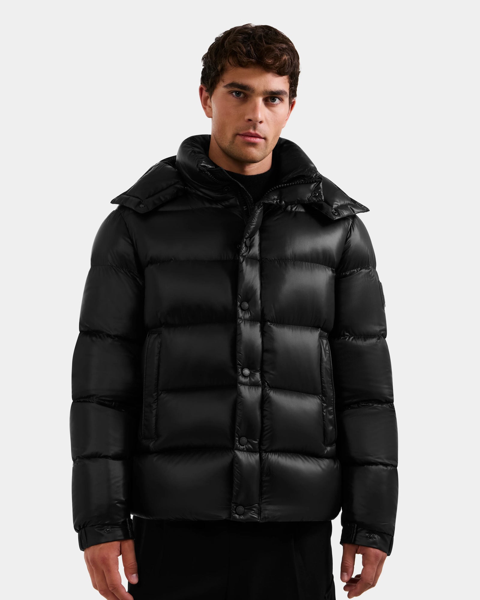 A man with short dark hair models the Rudsak BRAYDEN S - M125525 Black water-resistant quilted down puffer jacket with a high collar, standing against a plain light gray background.
