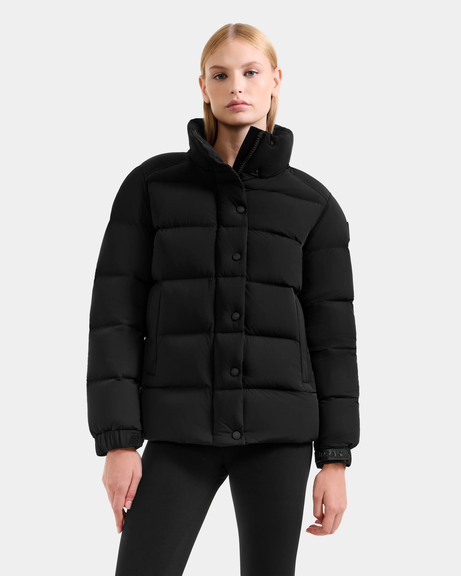 A woman with straight blonde hair confidently models the Rudsak BREE M - L125582 Black women’s down puffer jacket with a high collar and black leggings against a plain light gray background, showcasing this cold-weather essential.