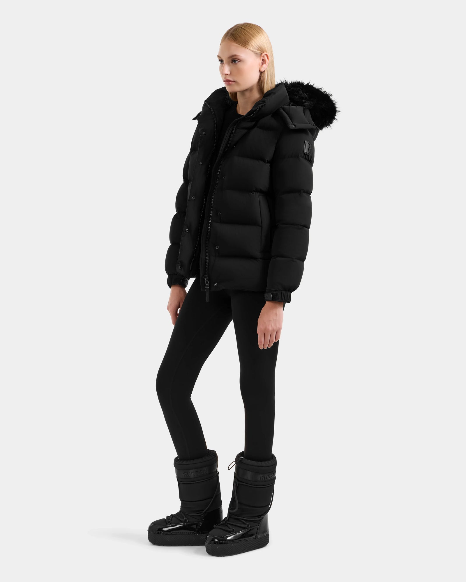 A woman wears the Rudsak BREE M - L125582 Black down puffer jacket with removable hood, paired with black leggings and chunky winter boots—an essential cold-weather look against a plain light background.