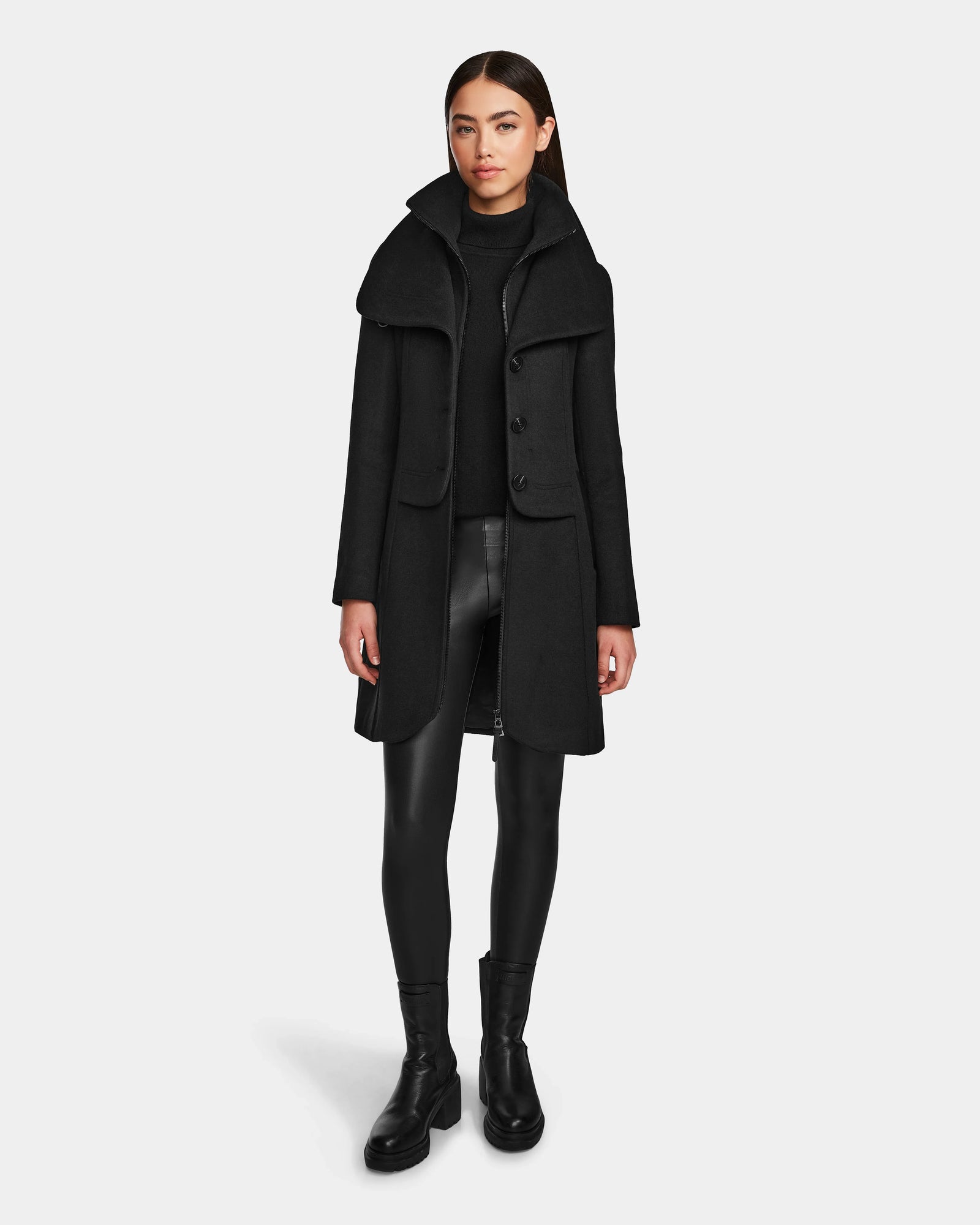 A woman with straight brown hair and a neutral expression stands against a plain background wearing the Rudsak CAROLINE - L124543 Black premium wool blend knee-length coat over a black turtleneck, vegan leather pants, and black ankle boots.