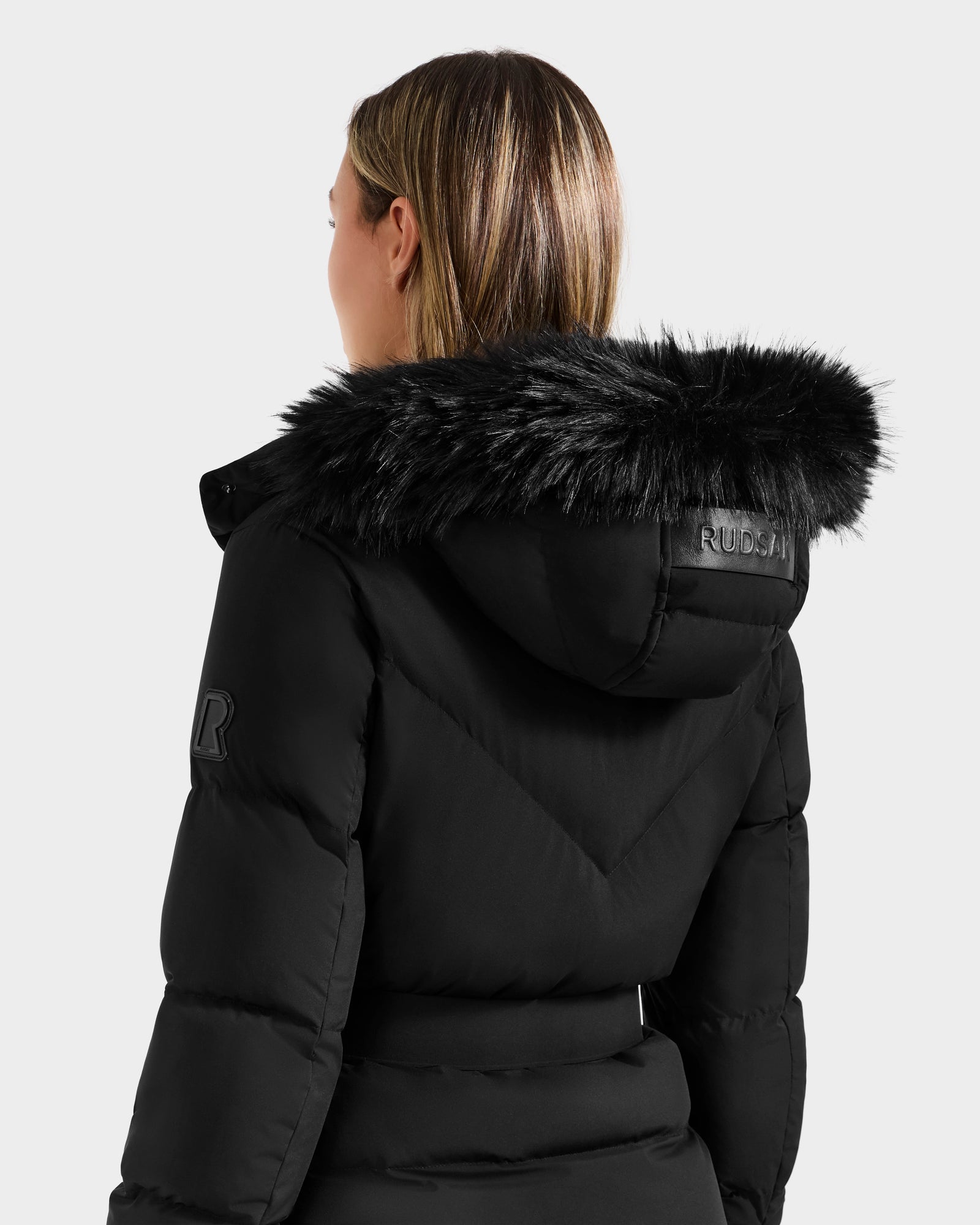 A person with light brown hair is seen from behind wearing the Rudsak CHRISTELLE M - L125542 Black weather-resistant quilted down puffer, featuring a black faux fur-trimmed hood and "RUBCAN" on the back of the hood.