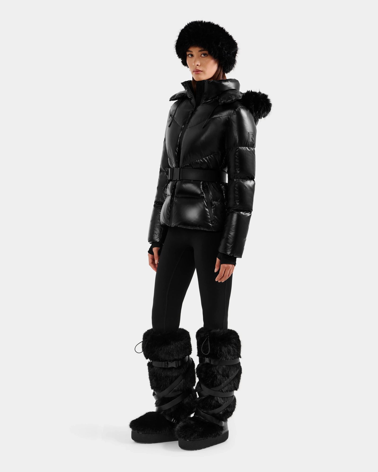 A woman wears the Rudsak CHRISTELLE S - L125544 Black down puffer jacket with a belt, paired with a black fur hat, fitted black pants, and tall faux fur-trimmed boots, standing against a plain white background.
