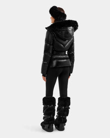 A person faces away wearing the Rudsak CHRISTELLE S - L125544 Black women’s down puffer jacket with faux fur trim, paired with fitted black pants, tall furry boots, and a fluffy headband for a chic, sporty winter look.