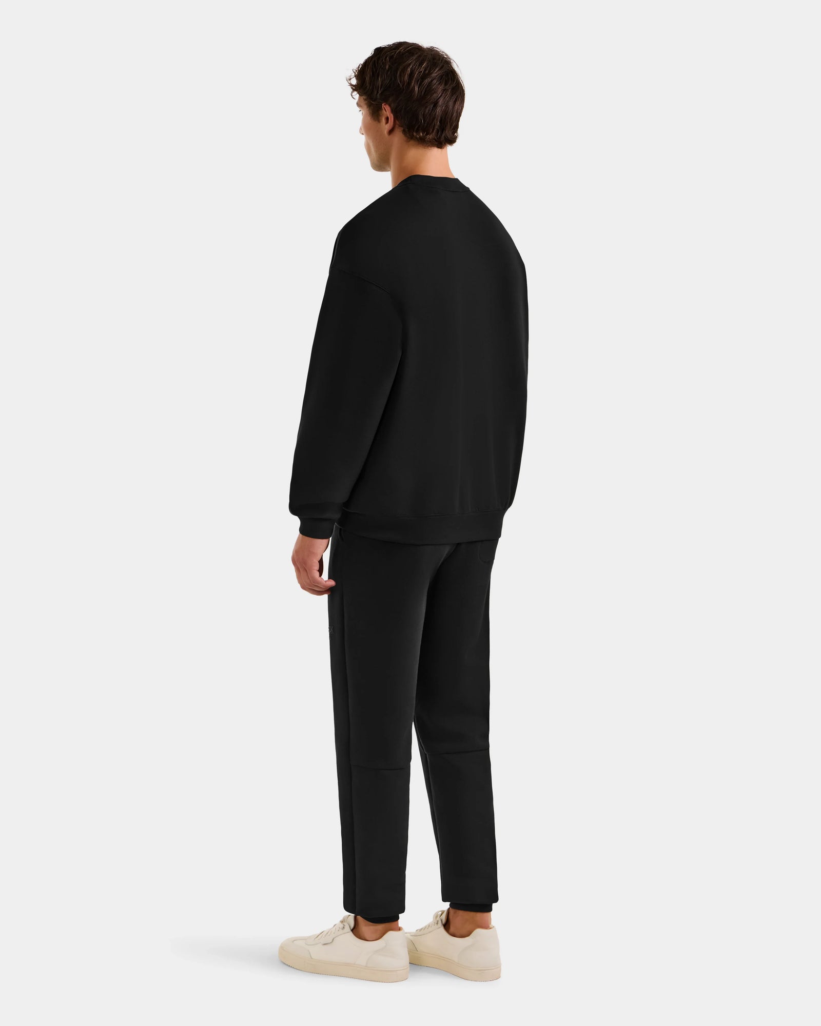 A man stands facing away, wearing the Rudsak COLSON - M525501 Black crewneck sweatshirt with matching black pants and white sneakers, set against a plain light gray background.