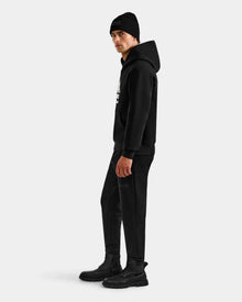 A person wearing the Rudsak DARWIN - M525502 Black hoodie, black beanie, pants, and boots stands sideways against a plain white background.