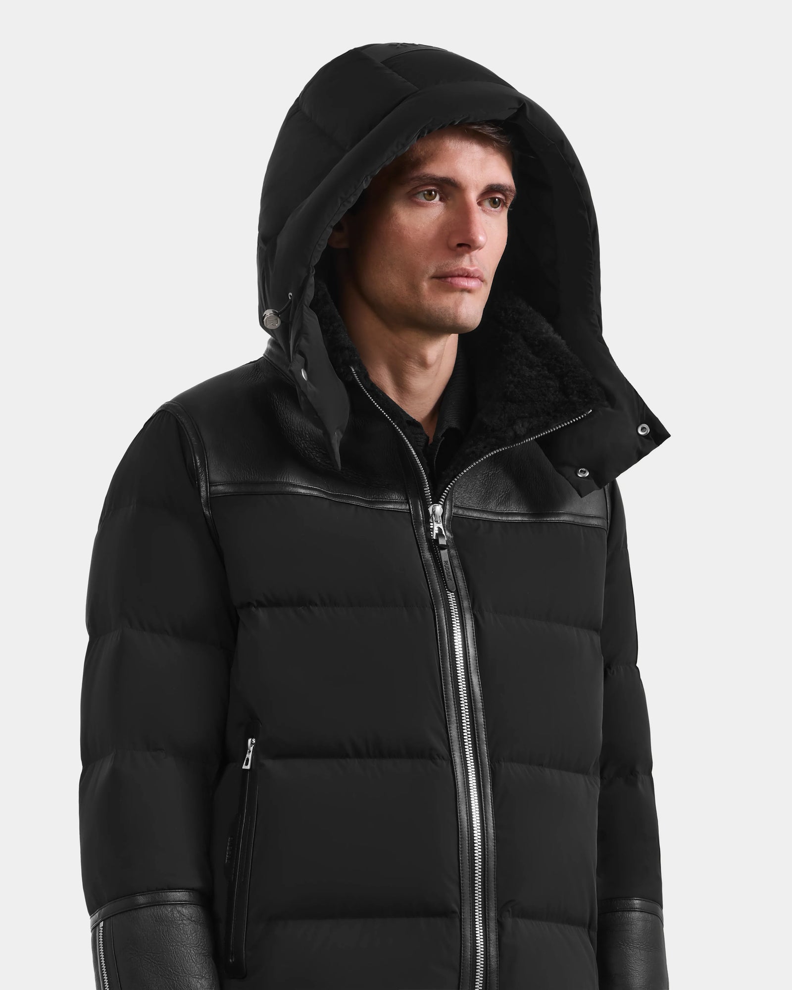 A man stands facing forward against a white background, wearing the Rudsak DAVID - M125508 Black, a men’s shearling down puffer coat with leather accents and hood up for a sleek, modern winter look.