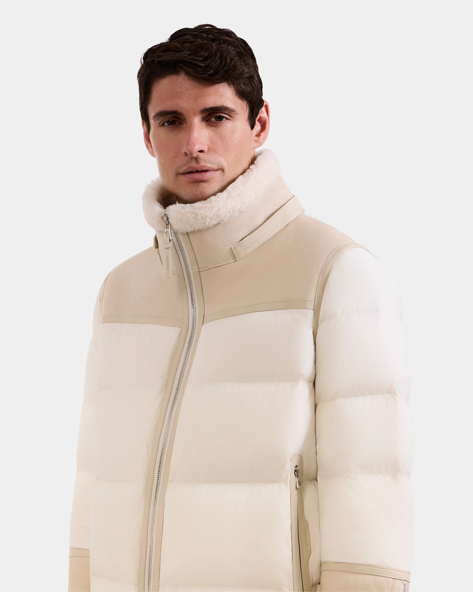 A man with short dark hair wears the Rudsak DAVID - M125508 Vanilla men’s shearling puffer jacket, featuring a high lamb shearling-lined collar and zipper front, standing against a plain white background.