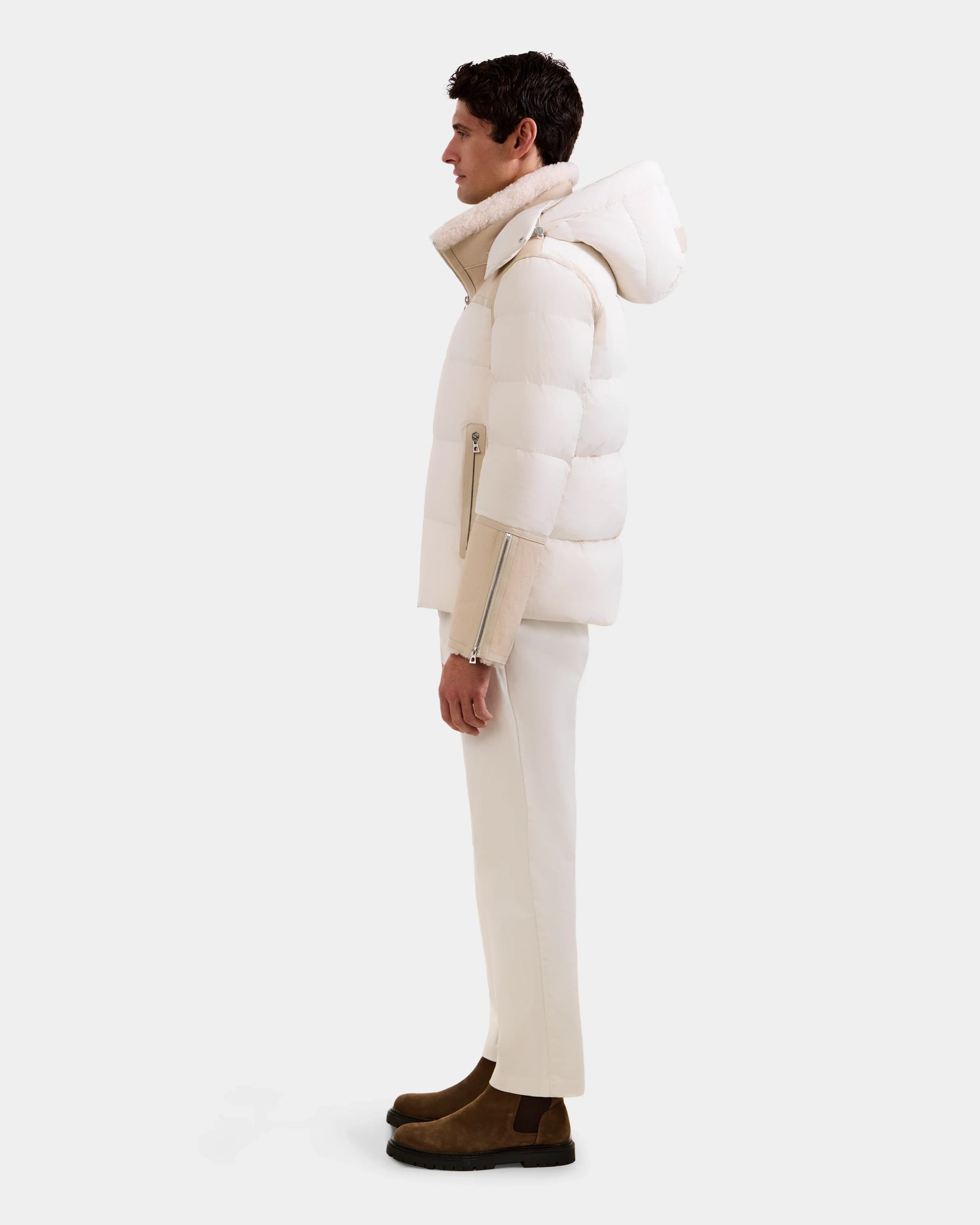 A man stands in profile against a plain background, wearing the Rudsak DAVID - M125508 Vanilla men's shearling puffer jacket with a hood, matching off-white pants, and brown suede boots.