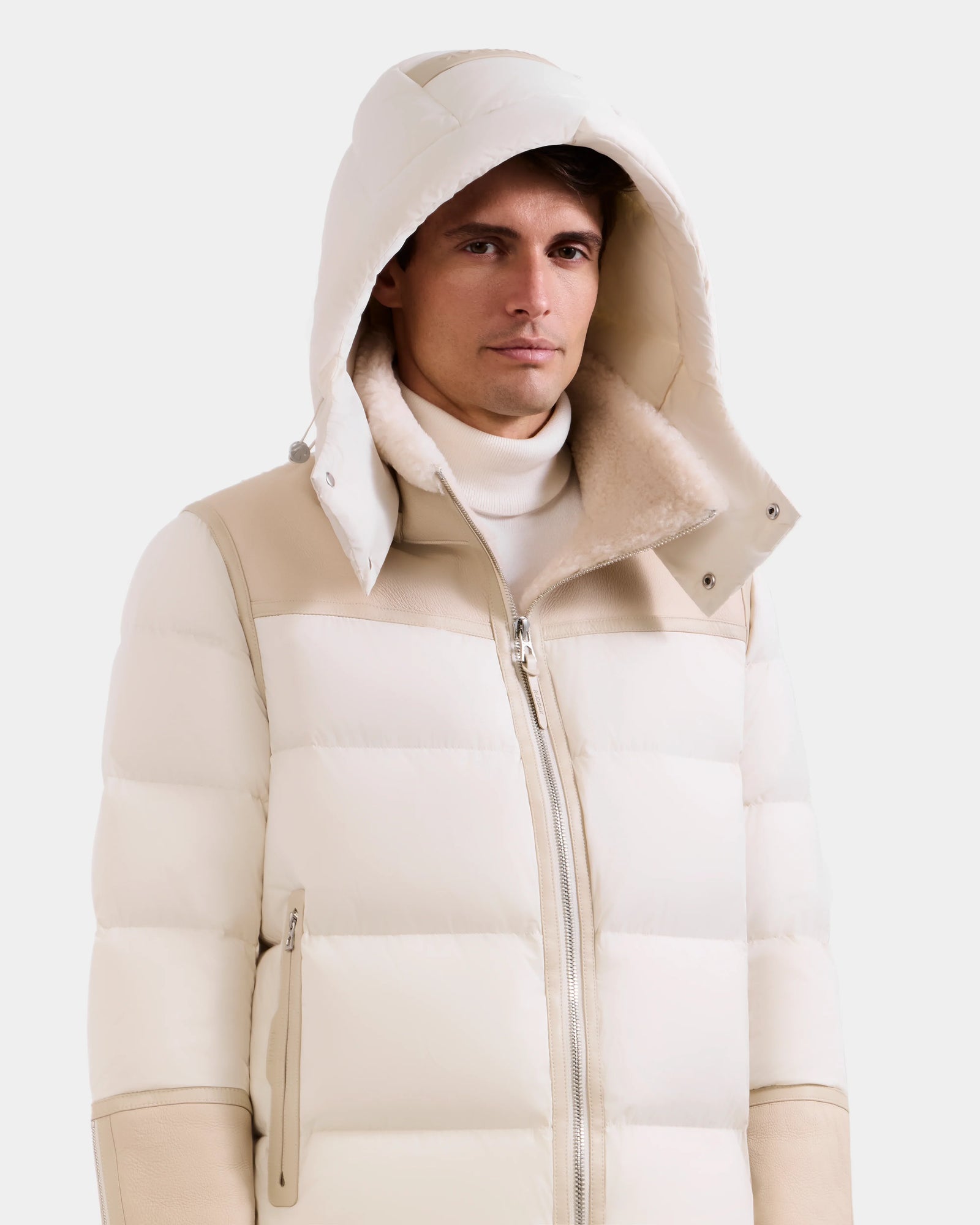 A man wears the Rudsak DAVID - M125508 Vanilla men's shearling puffer jacket with hood and high collar over a white turtleneck, standing against a plain light background.