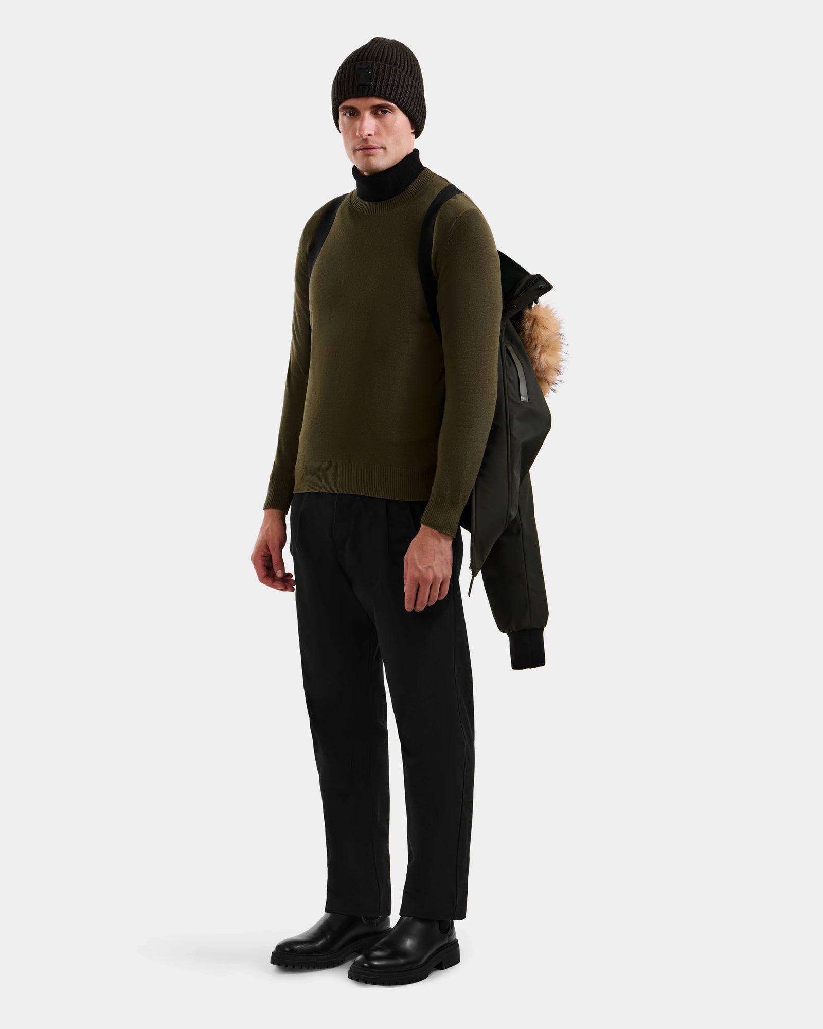 A man wearing the Rudsak DEACON - M123513 Dark Olive winter coat stands facing forward, dressed in black pants, boots, and a beanie. A bomber-style RDS-certified down jacket hangs from his black backpack against a plain white background.