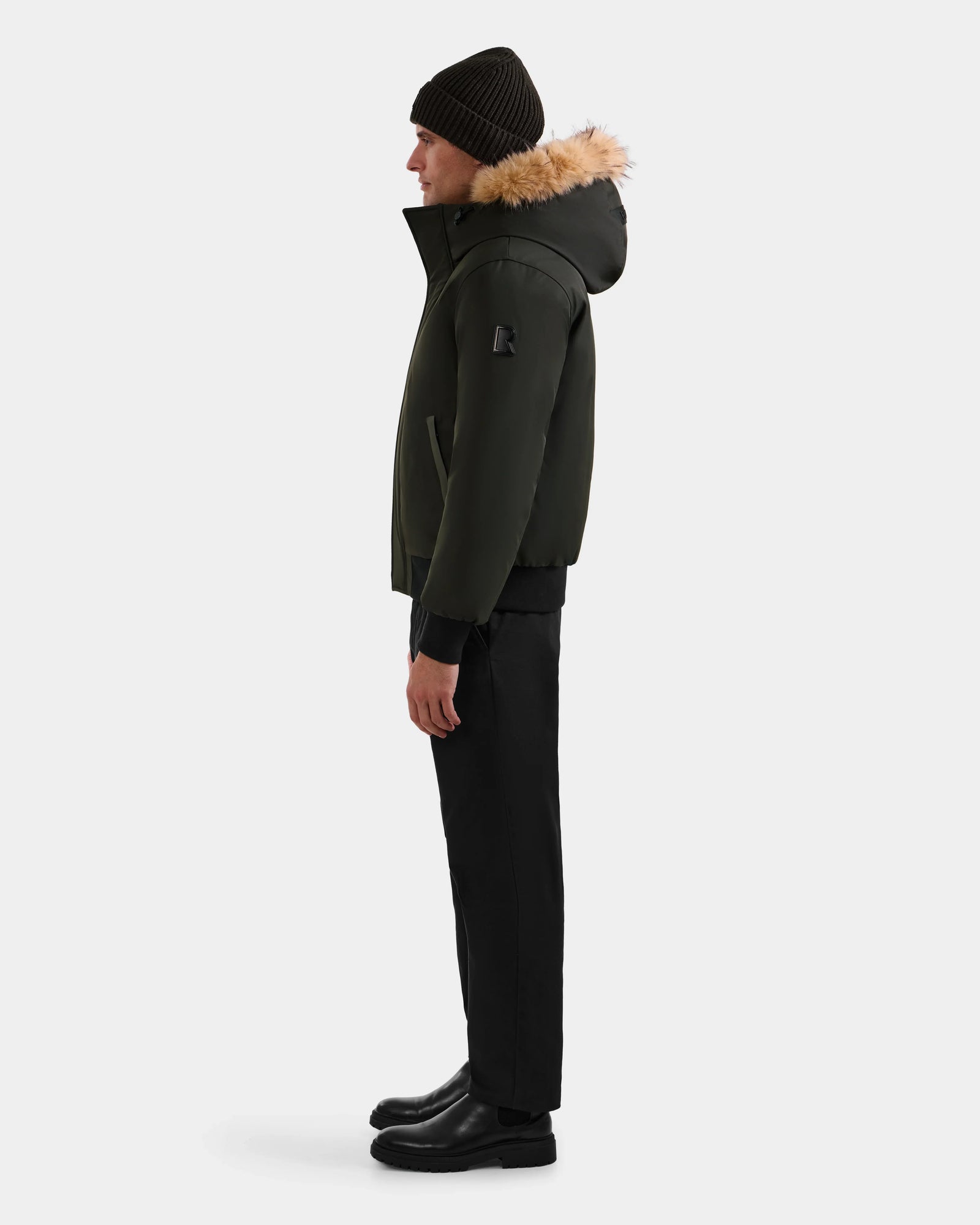 A person stands in profile against a white background wearing the Rudsak DEACON - M123513 Dark Olive men's bomber-style down puffer coat with fur trim, paired with black pants, boots, and a knit beanie.