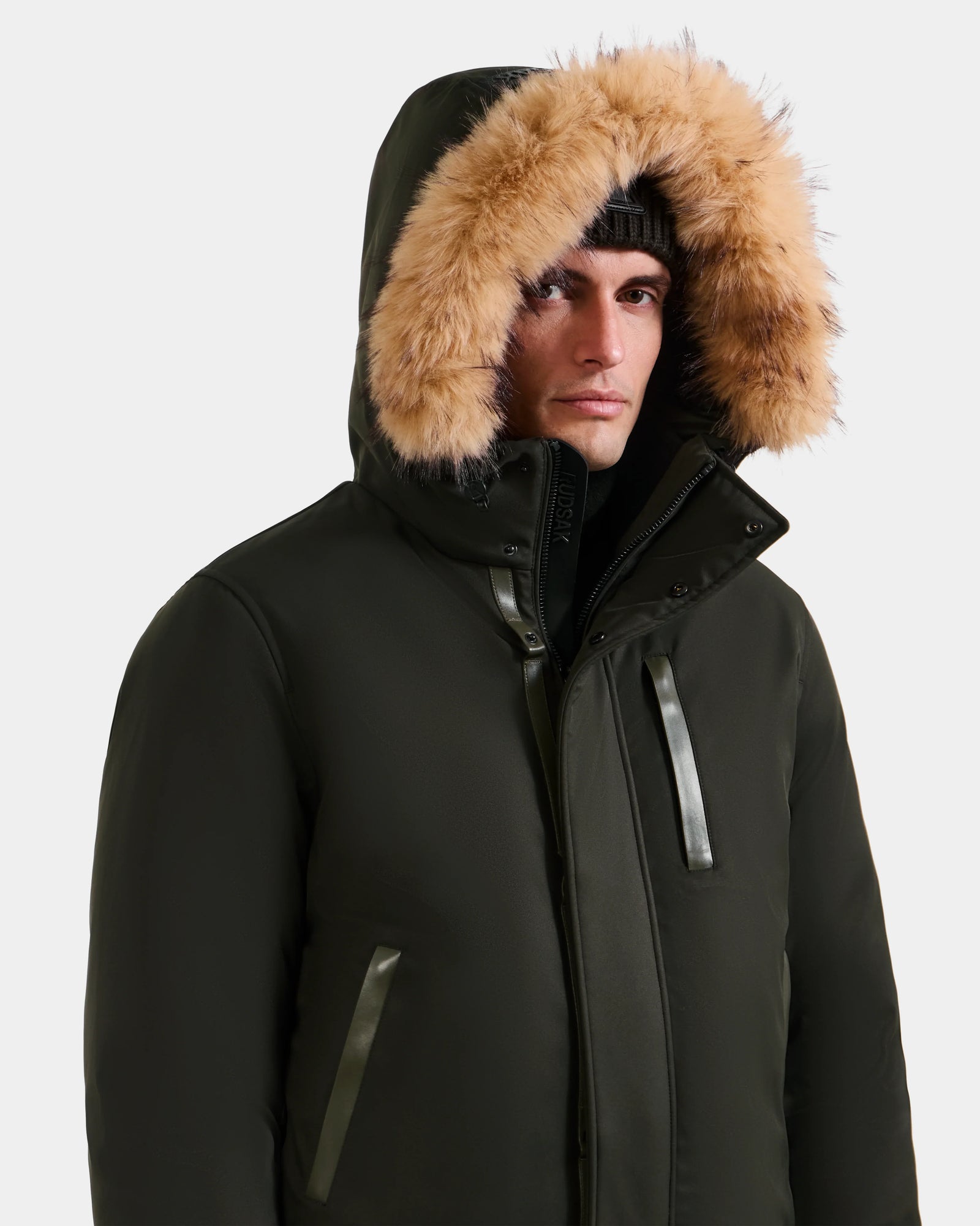 A person wearing the Rudsak DEACON - M123513 Dark Olive men's bomber-style down puffer with a fur-lined hood and a black knit beanie, looking forward against a plain light background.