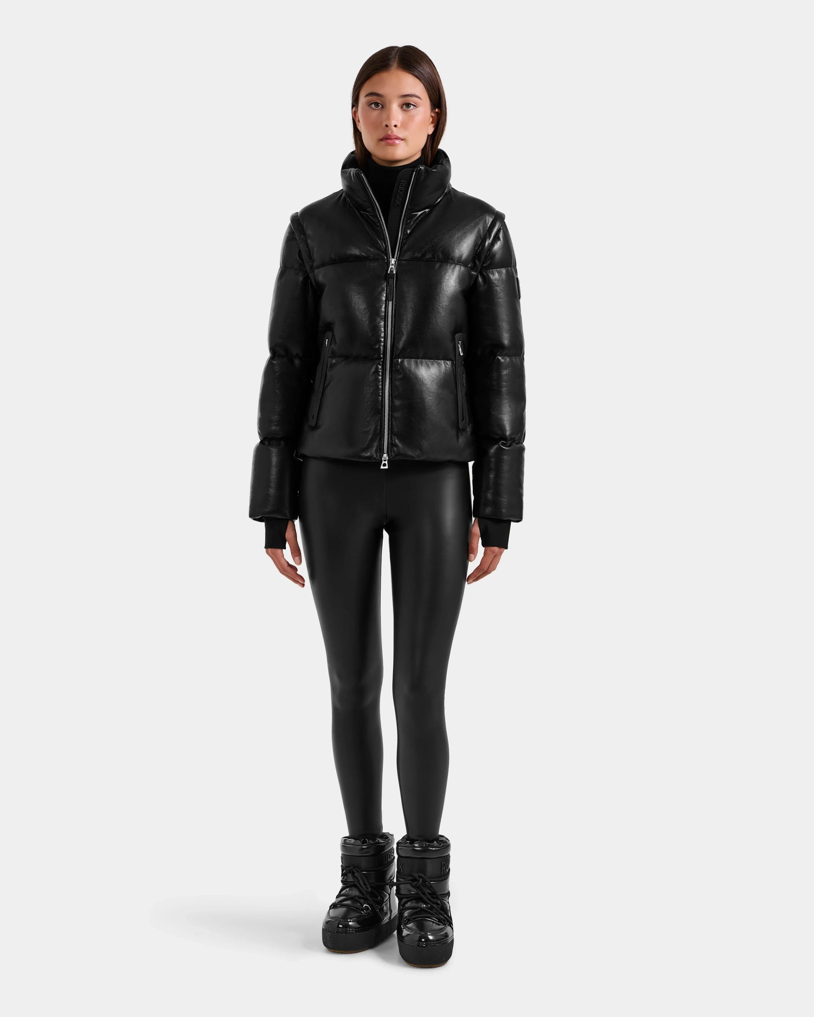 A woman with straight brown hair and a neutral expression models the Rudsak DELILAH PX - L125537 Black women’s vegan-leather puffer, paired with black leggings and winter boots, standing against a plain white background.