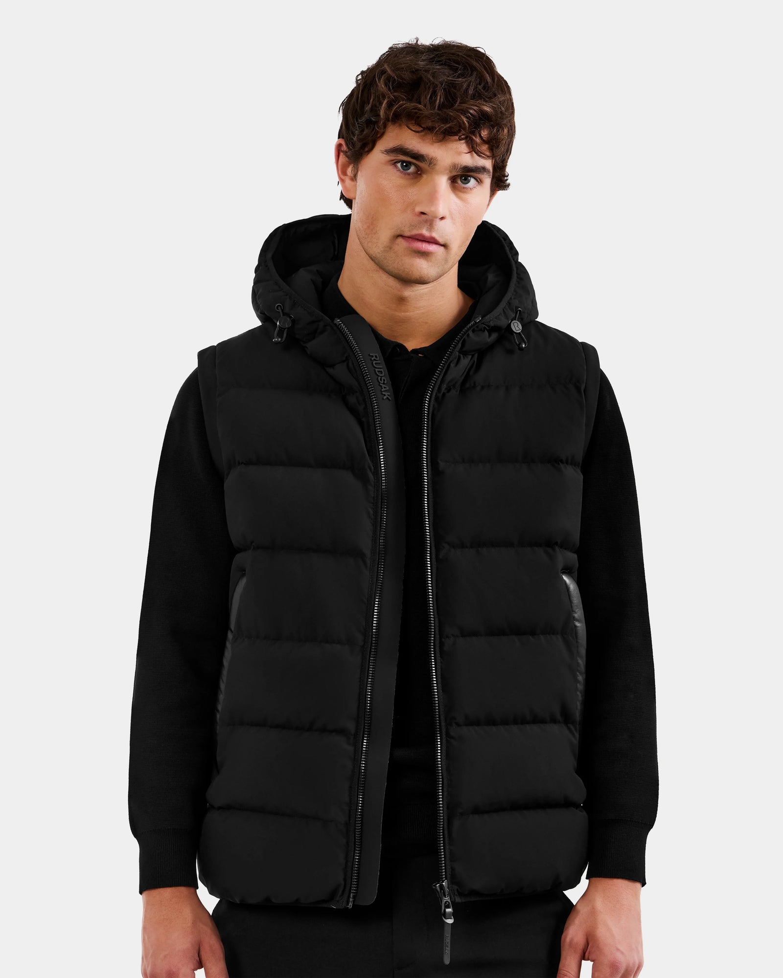 A young man with wavy brown hair wears the Rudsak DEMIAN M-R - M123504 Black men's down puffer vest over a black long-sleeve shirt, standing against a plain light gray background and facing forward with a neutral expression.