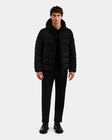 A man with short, curly brown hair stands against a plain background, facing forward with a neutral expression. He wears the Rudsak DEMIAN M-R - M123504 Black men's down puffer jacket, paired with black pants and black shoes.