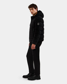 A man stands sideways against a plain light background, wearing the Rudsak DEMIAN M-R - M123504 Black men's down puffer jacket with black pants and black shoes.