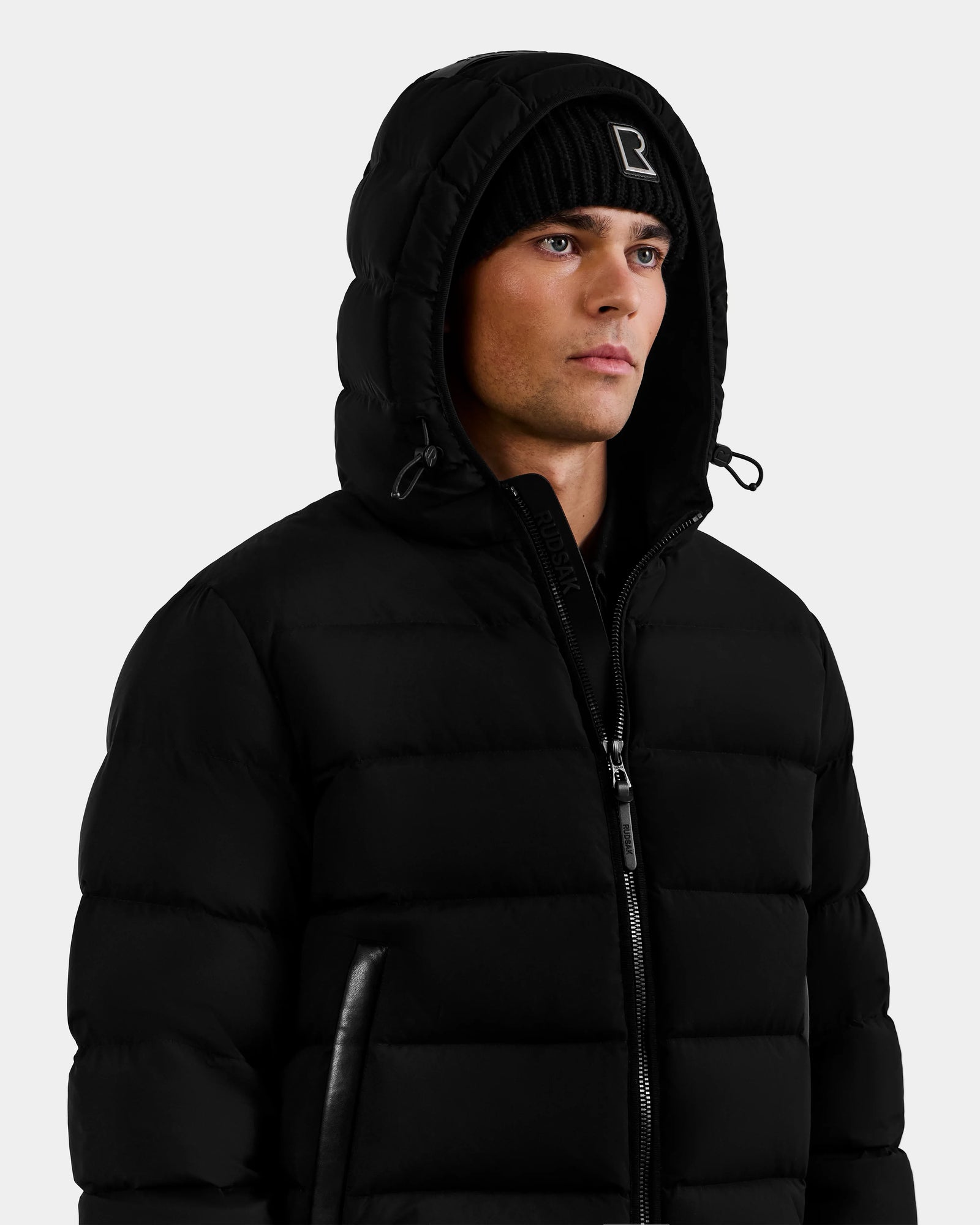 A man wearing the Rudsak DEMIAN M - M123507 Black down puffer jacket and a black beanie with a white "R", looking slightly to the side against a plain light background.