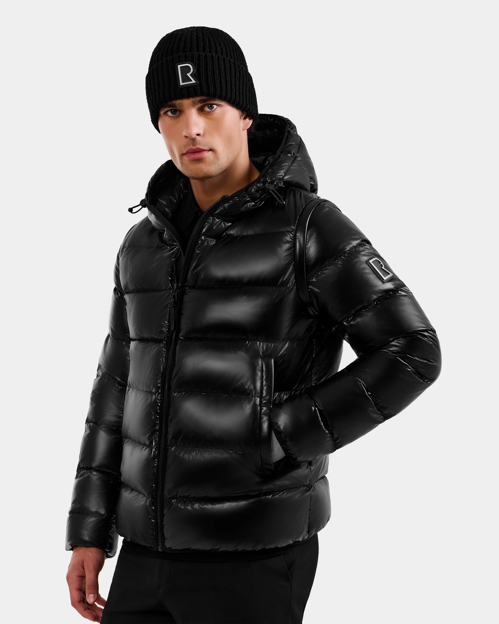 A man stands against a light gray background, wearing the Rudsak DEMIAN S-R - M123505 Black men's down puffer jacket with a hood and a black beanie, one hand in his pocket, looking at the camera.