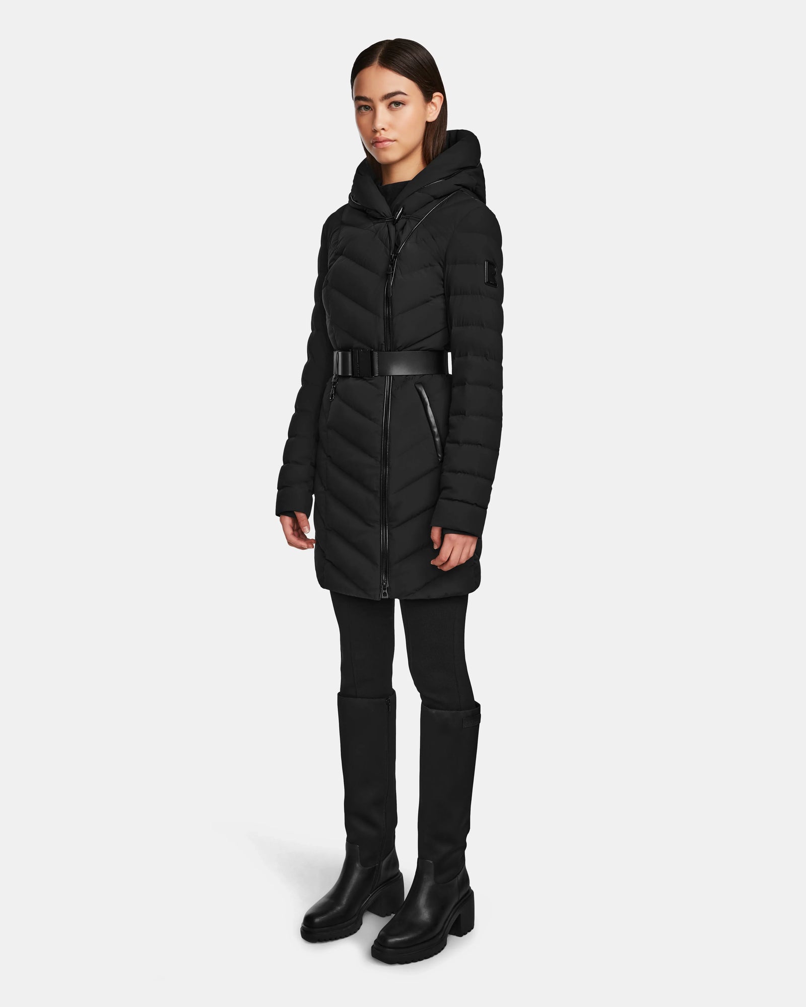 A woman stands facing forward against a plain light background, wearing the Rudsak DITA - L124556 Black puffer jacket with hood and belted waist, paired with black leggings and knee-high black boots.