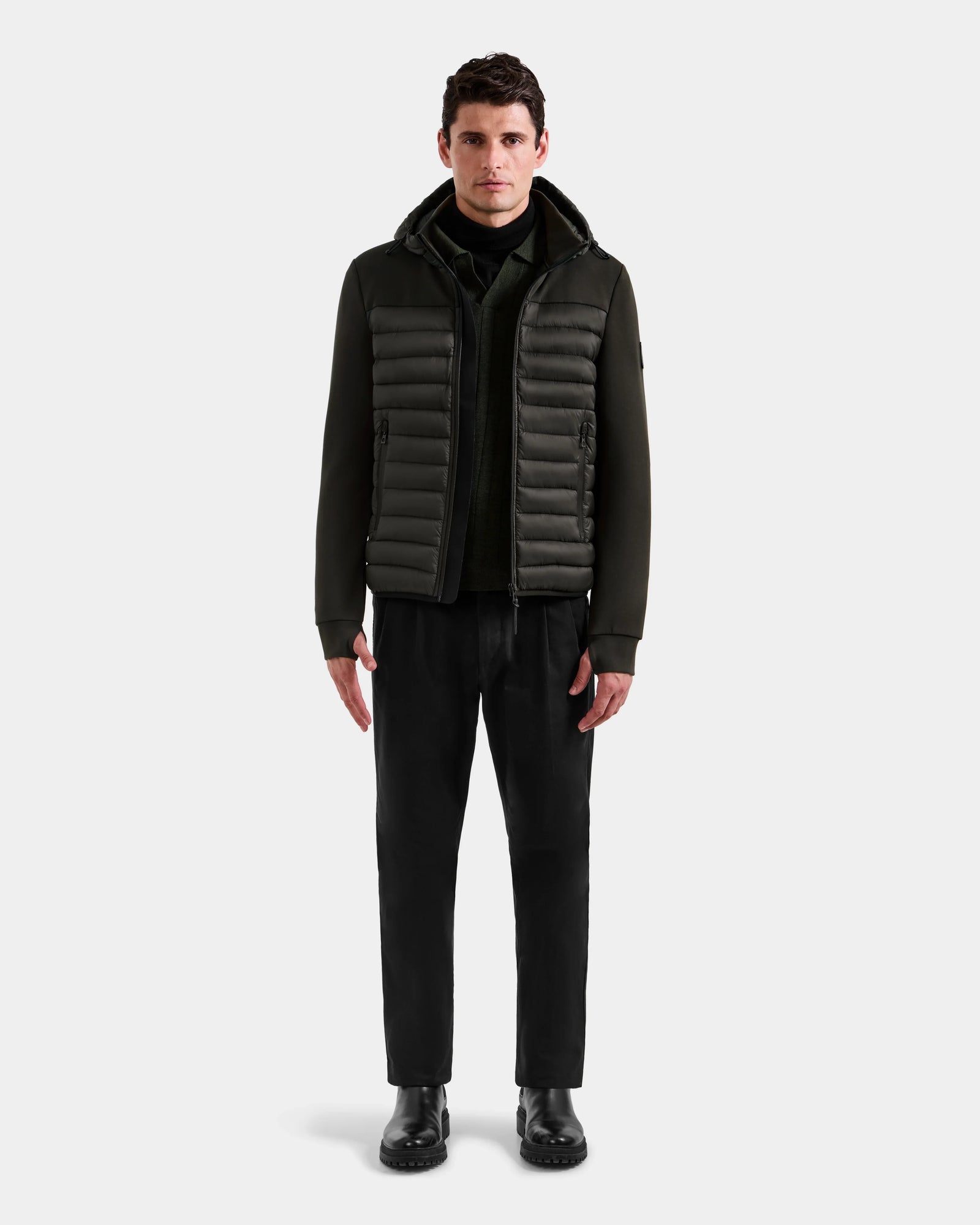 A man faces forward wearing the Rudsak DURIN M – M125530 Dark Olive men’s hooded jacket with lightweight down insulation and waterproof zippers, paired with black pants and boots, against a plain light gray background.