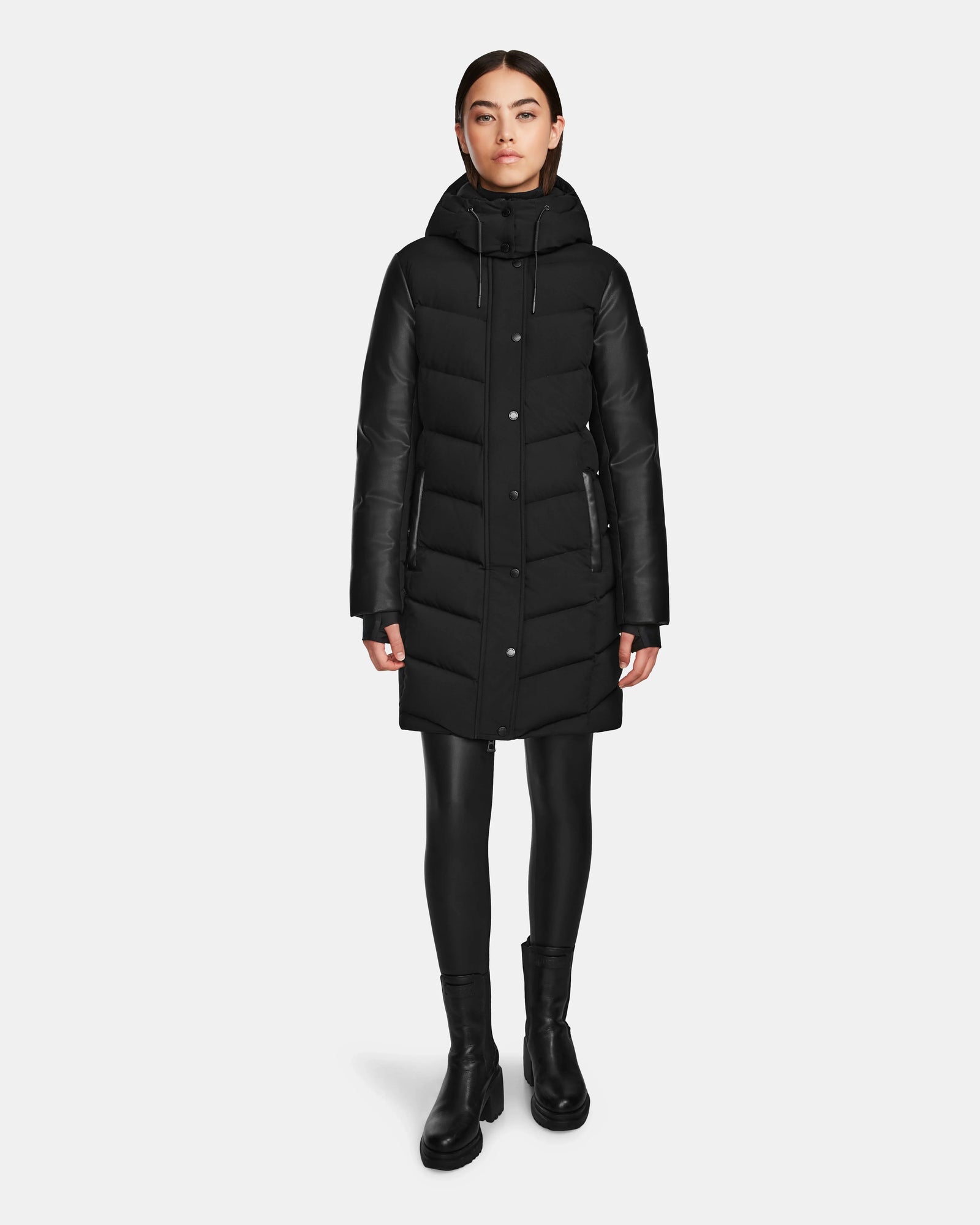 A woman stands facing forward against a plain white background, wearing the Rudsak ELSA - L123535 Black quilted down puffer with a hood, black leggings, and black ankle boots.