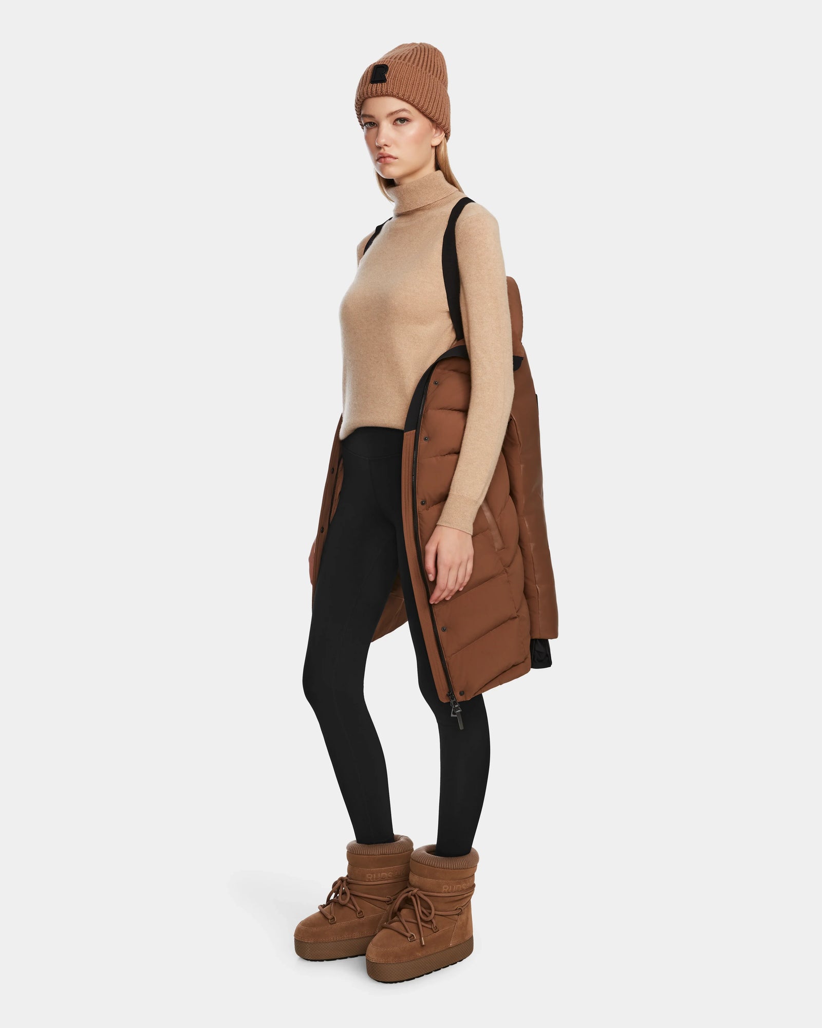 A woman stands against a white background in black leggings, a tan sleeveless turtleneck, and the Rudsak ELSA - L123535 Taupe quilted down puffer coat draped over one shoulder, styled with a tan knit beanie and brown winter boots.