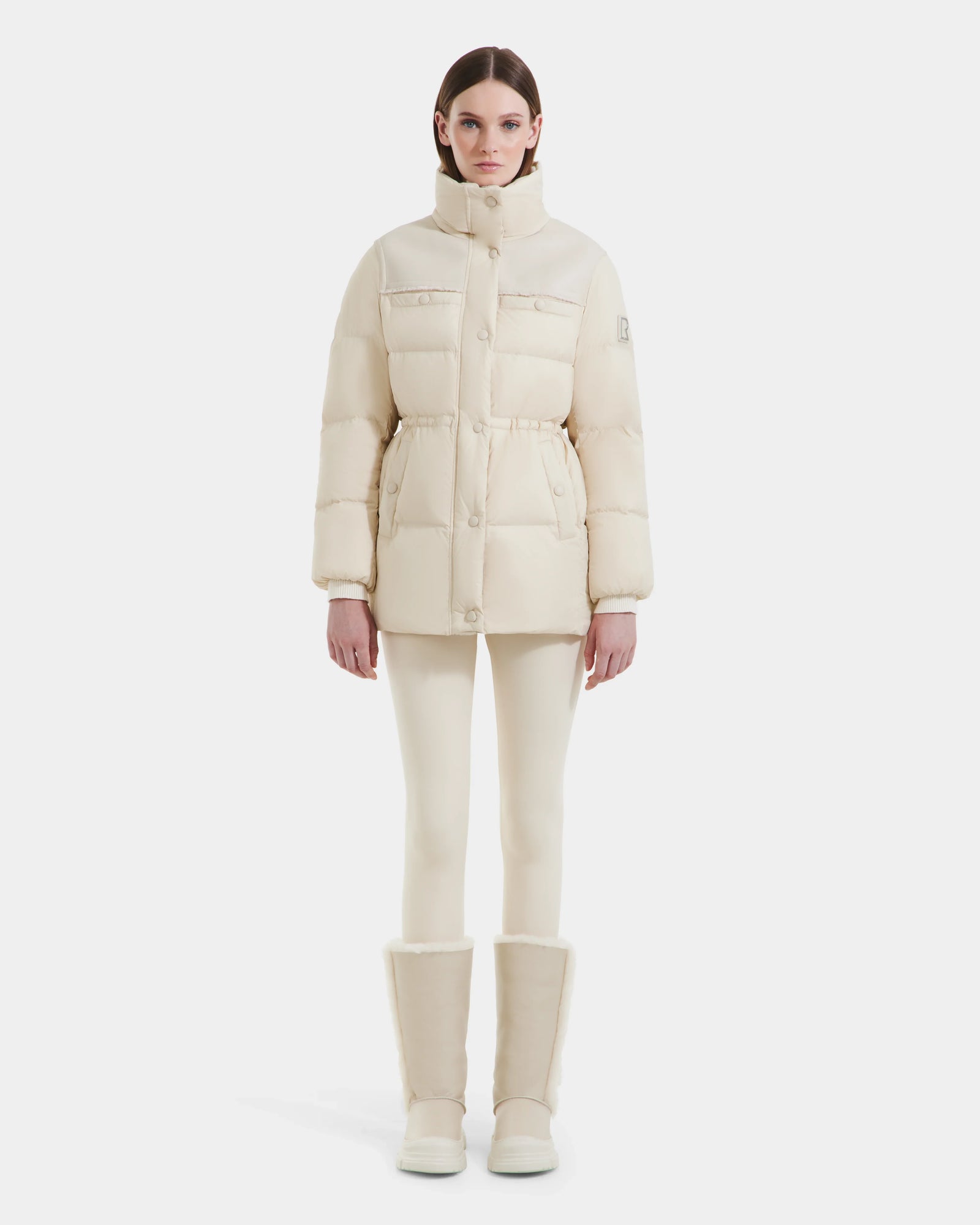 A woman with straight brown hair and a neutral expression faces forward, wearing the Rudsak EMBER M-M - L125524 Vanilla cream down puffer jacket, matching leggings, and tall boots against a plain white background.