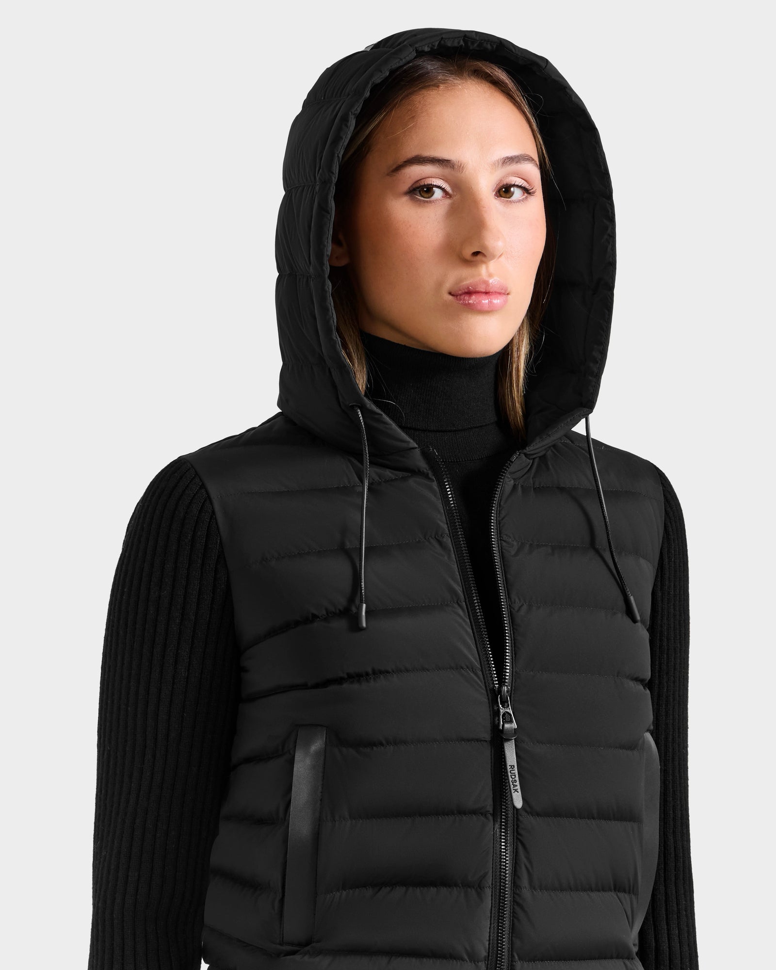 A woman wears the Rudsak EMILY - L125515 Black RDS-certified down quilted hooded vest over a ribbed turtleneck, standing against a plain light background and looking slightly to the side with a neutral expression.