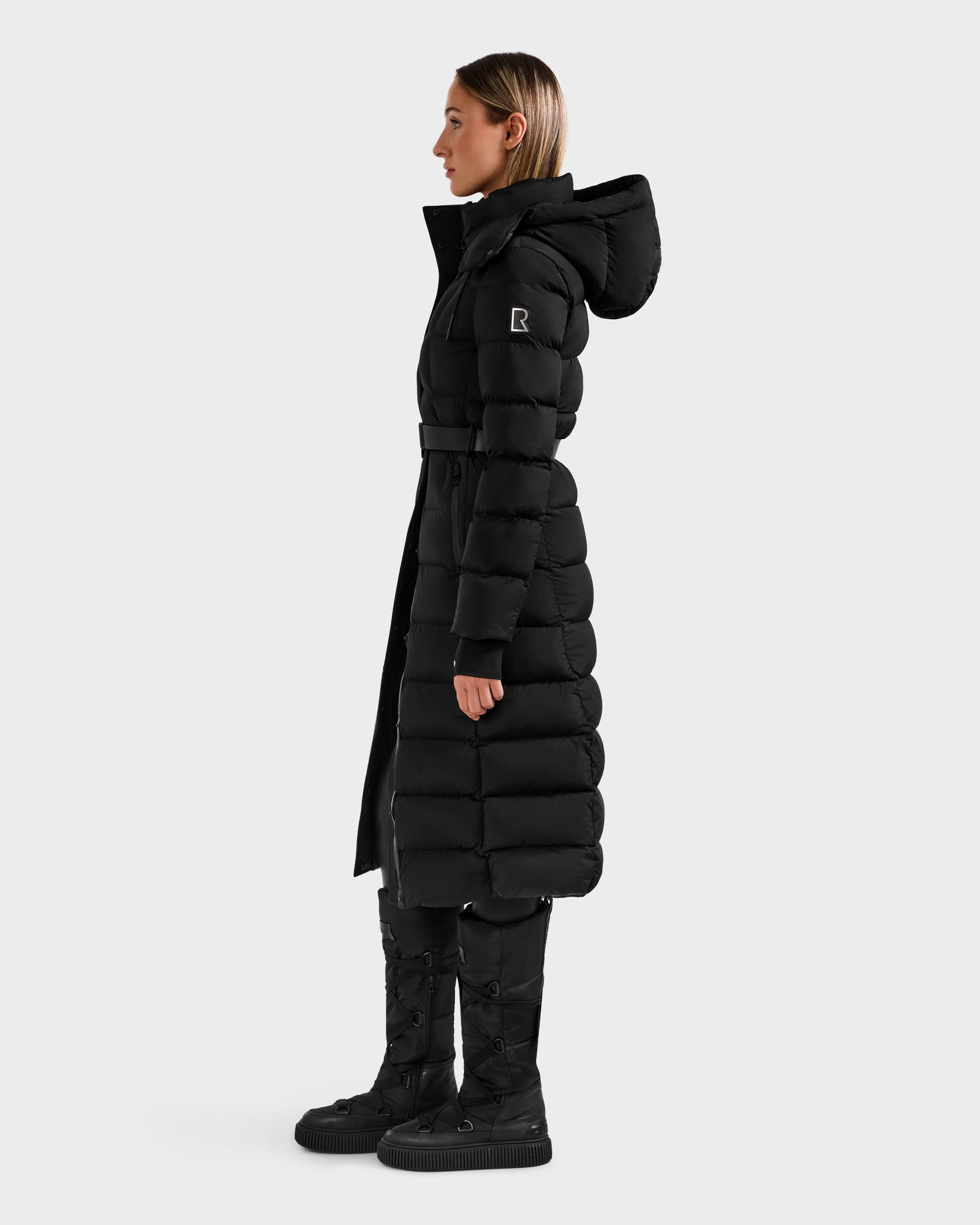 A woman in profile wears the Rudsak FRIDA M-L - L125608 Black long down puffer with a belted waist, sleeve patch, detachable hood, and matching black boots. The black coat stands out against a plain white background.