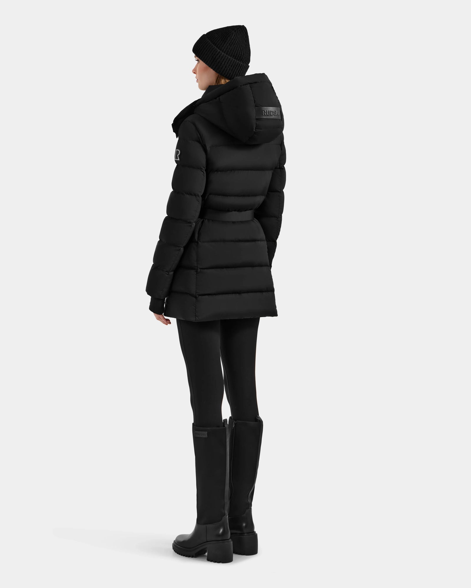 Dressed in the Rudsak FRIDA M-M - L125607 Black quilted down puffer with detachable hood, a black knit hat, black leggings, and knee-high boots, a woman stands facing away against a plain light background.