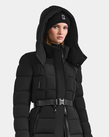 A woman in the Rudsak FRIDA M-M - L125607 Black quilted down puffer coat with detachable hood, belted waist, and a black knit logo beanie stands against a plain white background.