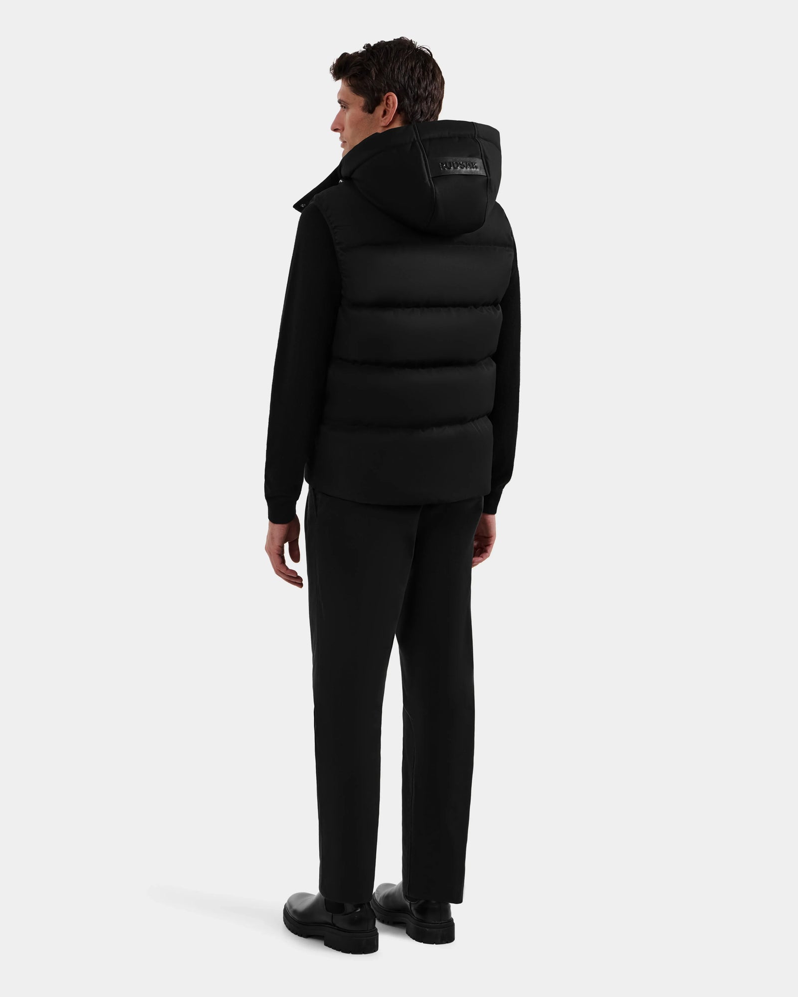A man faces away, wearing the Rudsak HARLOW - M125528 Black men’s puffer vest with removable hood, paired with black pants and shoes, set against a plain light background.