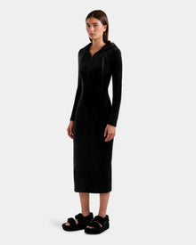 A woman with long straight hair and a neutral expression stands against a plain background wearing the Rudsak ISABELLA - L525502 black velour hooded midi dress with zipper front, paired with black fuzzy sandals.
