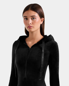 A young woman with straight brown hair wears the Rudsak ISABELLA - L525502 black zip-up hoodie with a hood and drawstrings, standing against a plain light gray background while looking directly at the camera with a neutral expression.